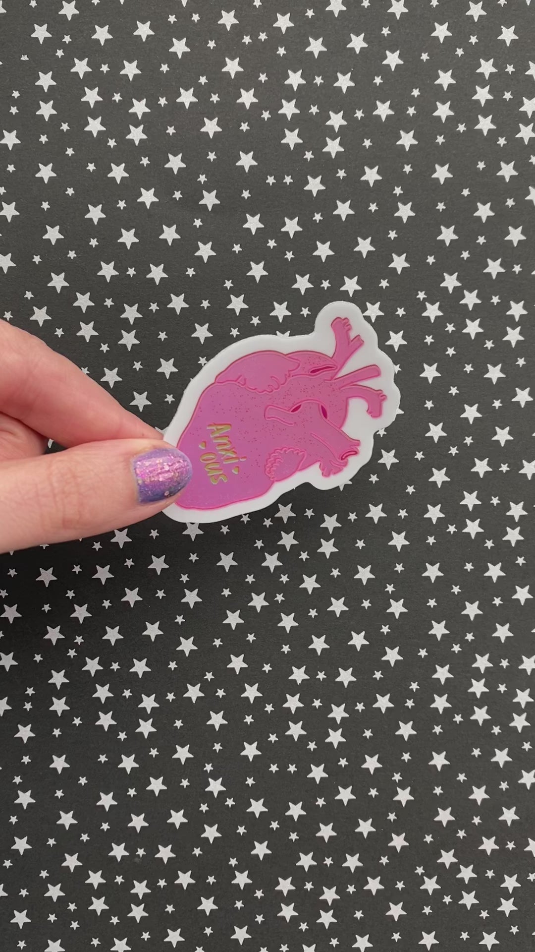 A person with purple nail polish holds a metallic sticker with a realistic human heart illustration and the the word "anxious" flanked by hearts.