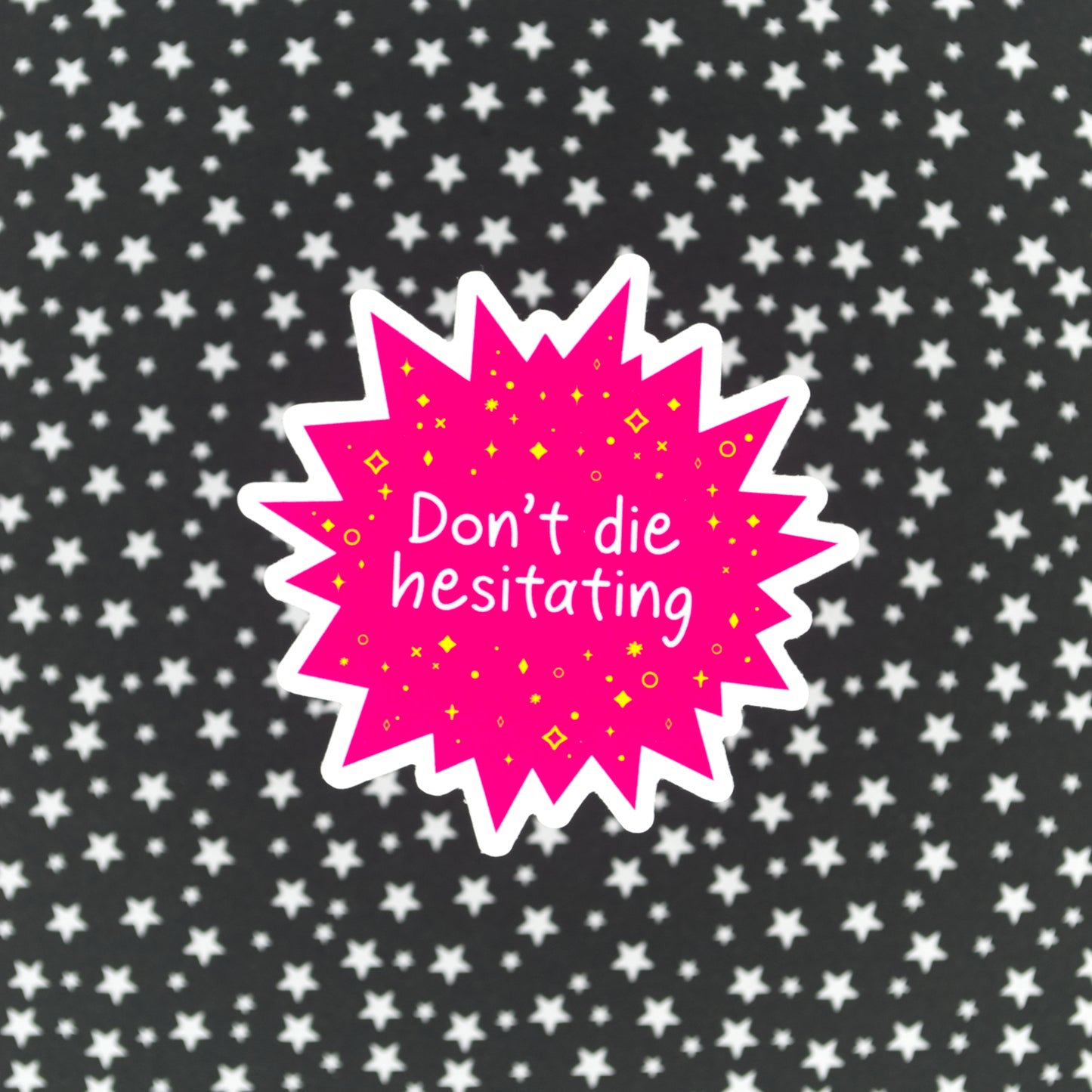 A bright pink star-burst shaped sticker with the words "Don't die hesitating" in white handwriting surrounded by yellow stars and sparkles. The sticker sits against a black background with silver stars