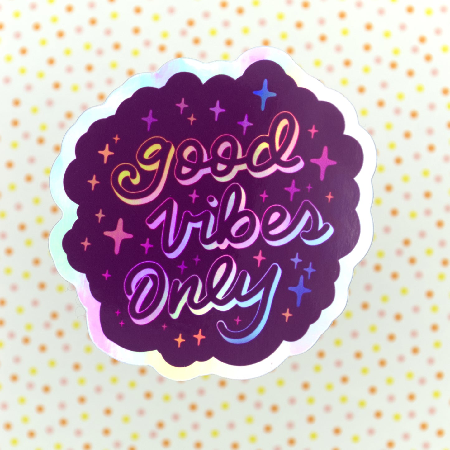A purple holographic sticker with the words "good vibes only" written in calligraphy and surrounded by rainbow sparkles. The sticker sits on a pinnk, yellow and orange spotted background.