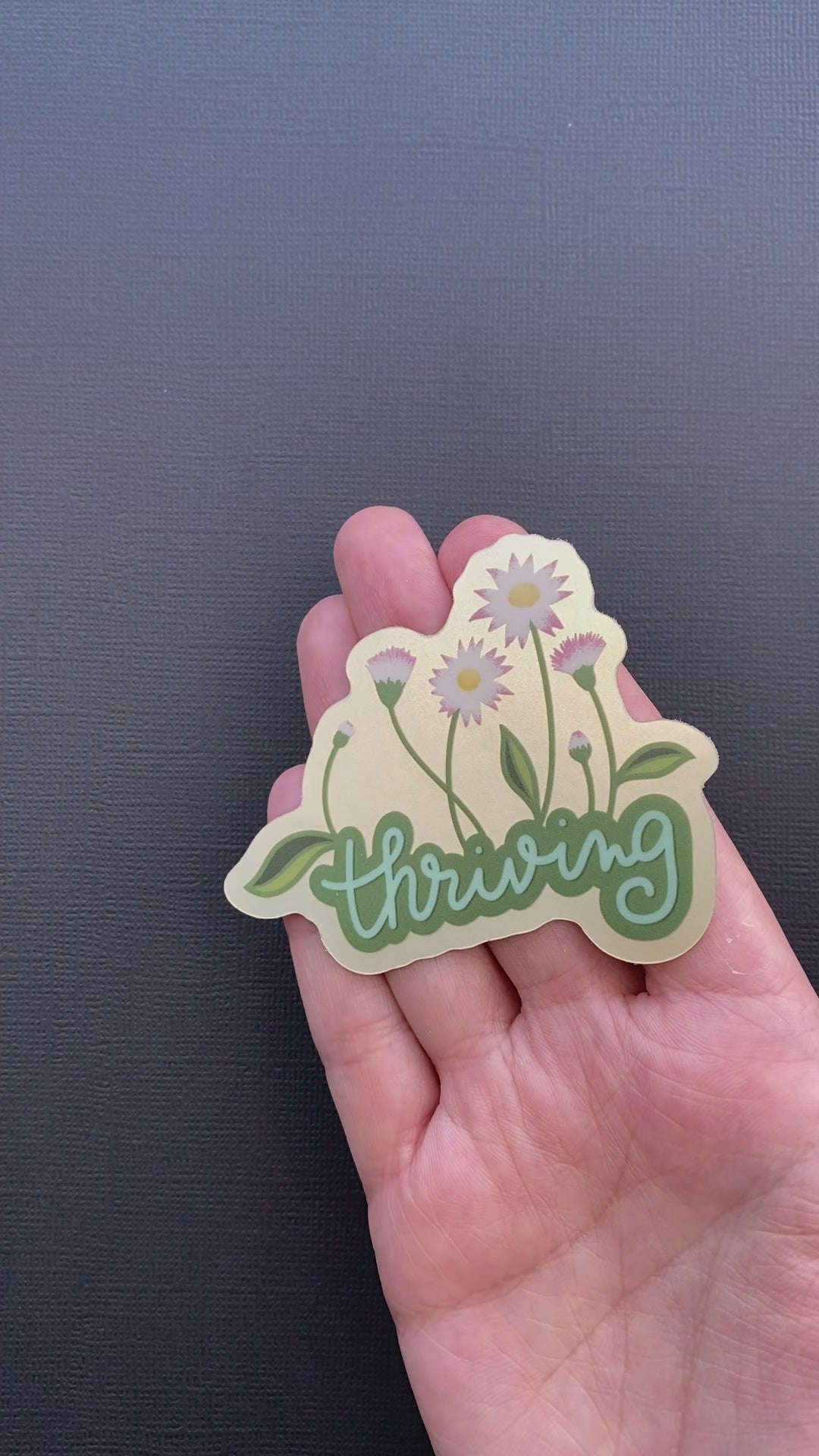 A video of a person's hand rotating the Thriving sticker so you can see the metallic effect.