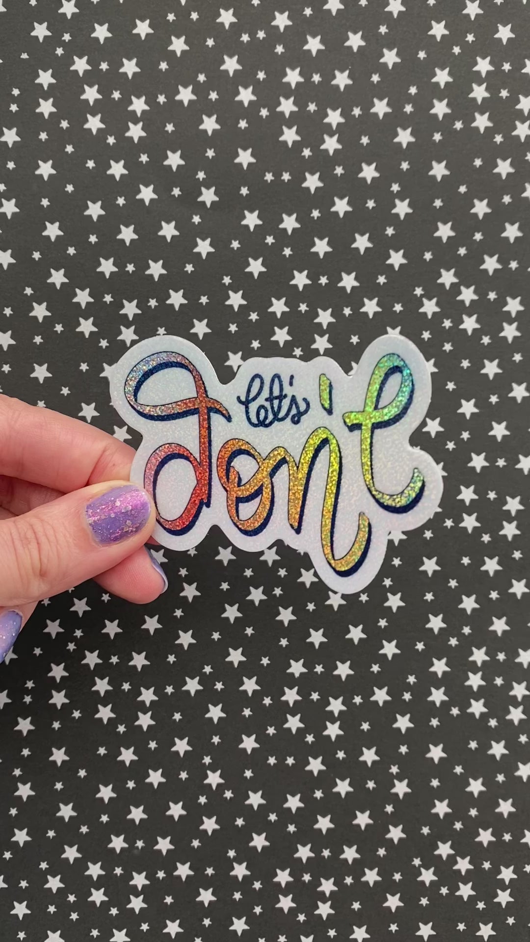 A person with purple nail polish holding a rainbow glitter sticker to show how the light hits the sticker. The sticker is held againt a black background with silver stars.