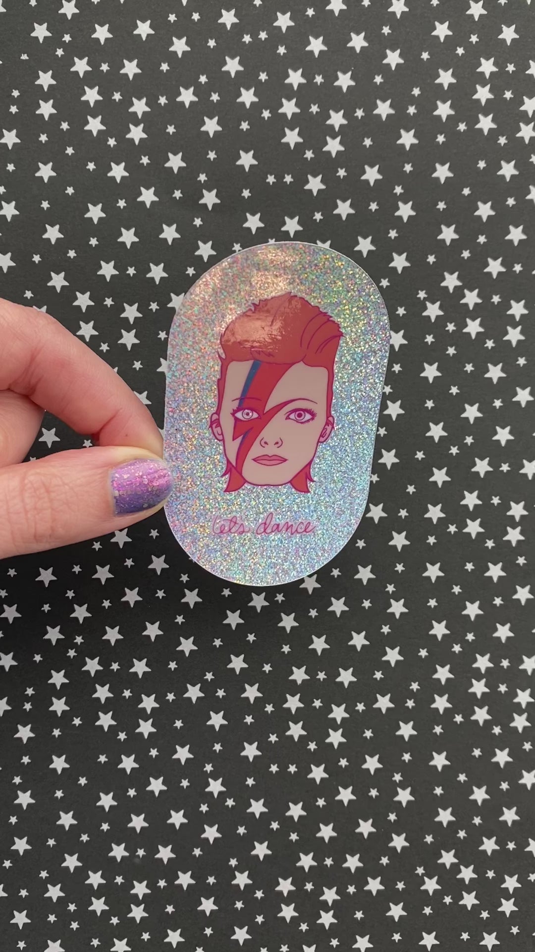 A person with purple nail polish holds a sticker featuring an illustration of David Bowie's head with the phrase "let's dance" on irridescent glitter vinyl. The sticker catches the light as the person turns it.