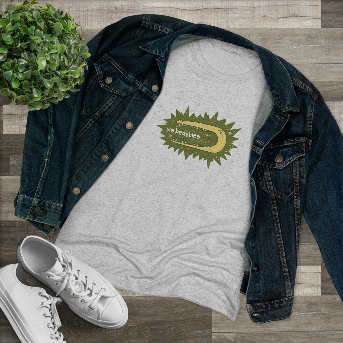 A varigated gray t-shirt with the We Beasties logo (a yellow alligator on a green star burst surround by pink stars and the words "We Beasties" in the alligator's mouth.) The shirt is shown with a demin jacket, white sneakers, and a plant against a wood floor.