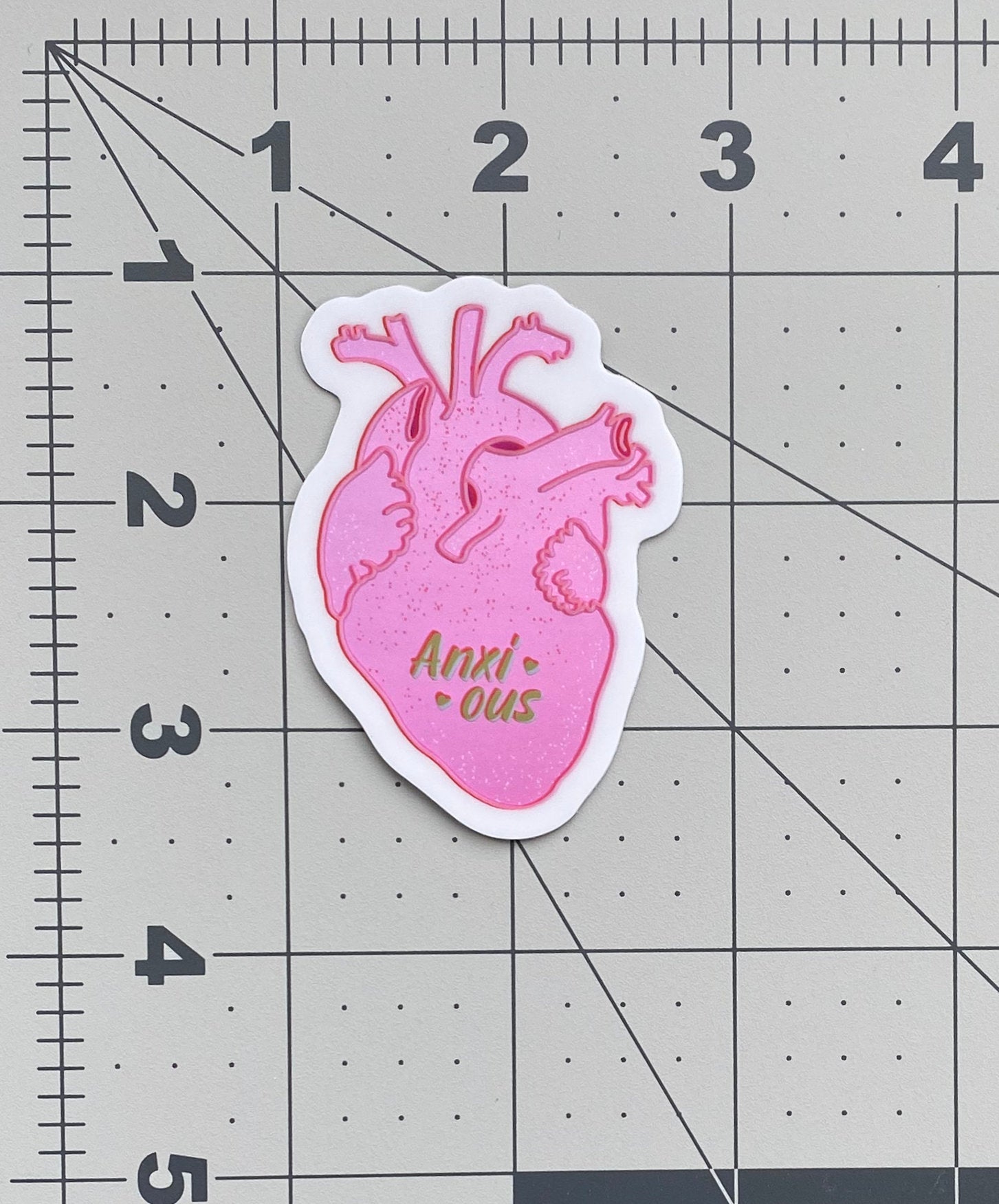 A metallic sticker with an illustration of a realistic human heart and the the word "anxious" flanked by hearts. The sticker sits on a ruler to show its size.