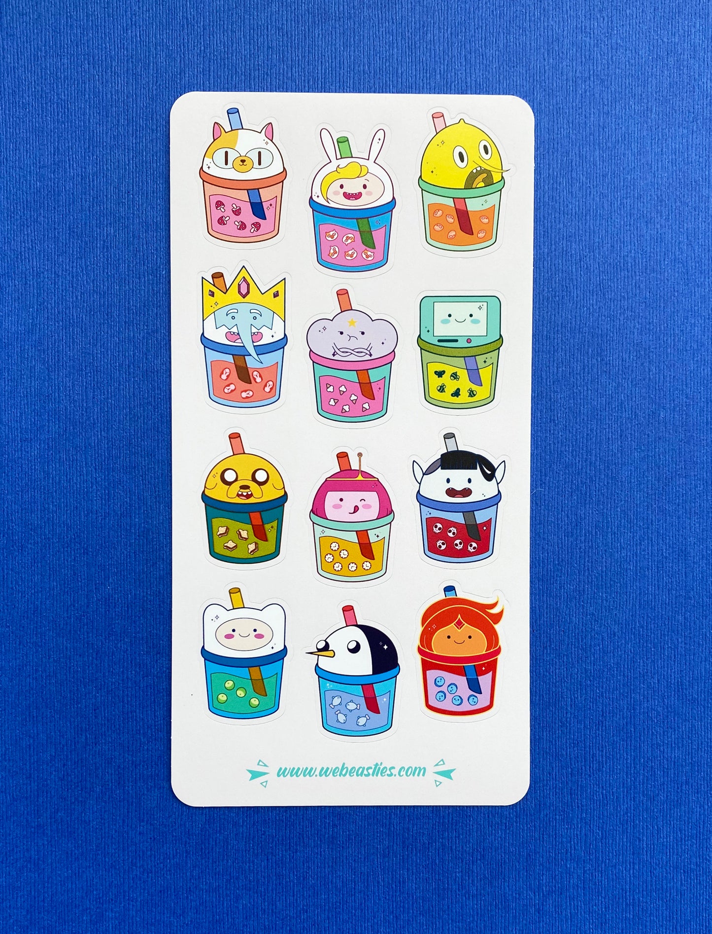 A photo of a sticker sheet with illustrations of 12 Adventure Time characters as bubble tea cups with lids, straws, and themed boba. The sticker sheet sits on a blue paper background.