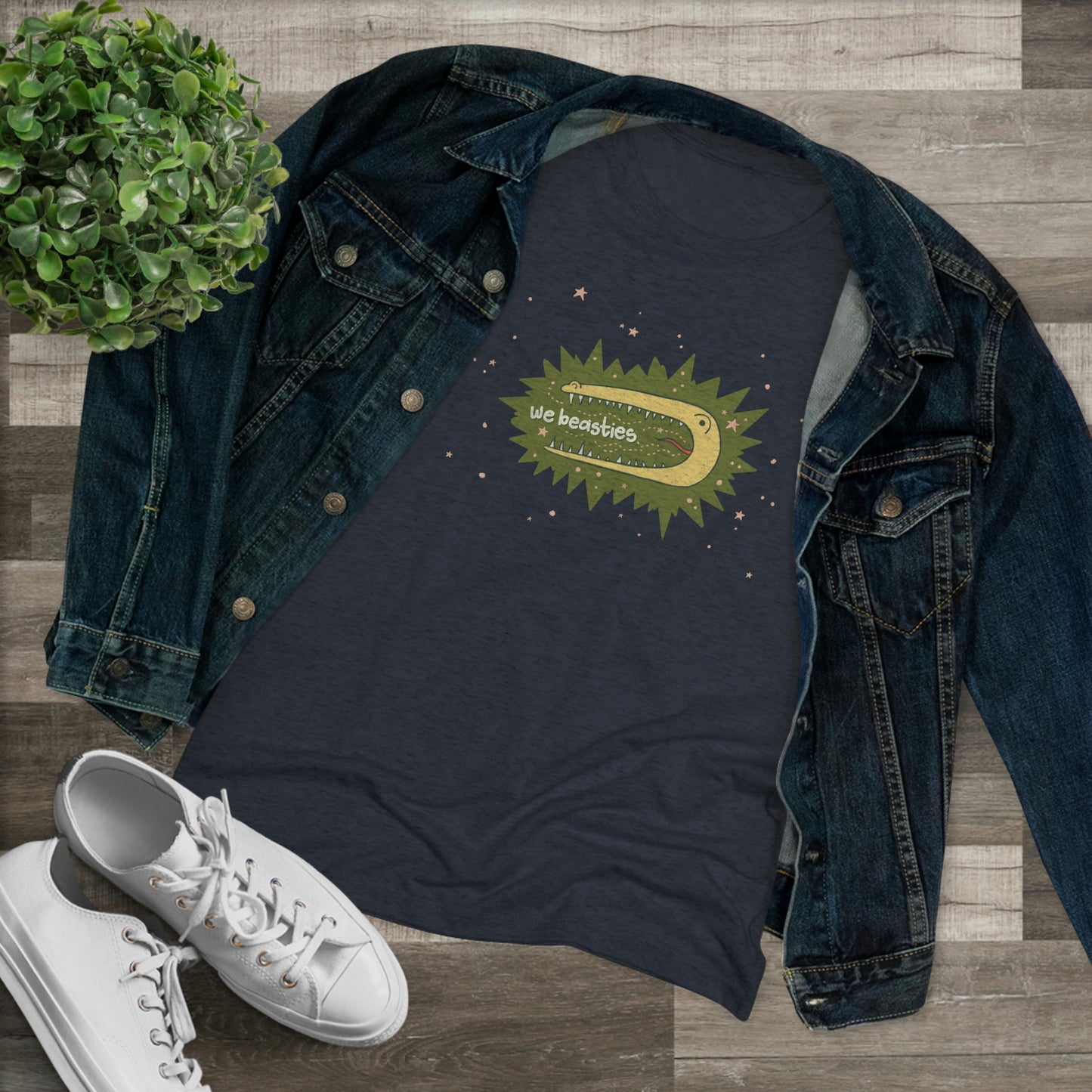 A varigated dark blue t-shirt with the We Beasties logo (a yellow alligator on a green star burst surround by pink stars and the words "We Beasties" in the alligator's mouth.) The shirt is paired with a denim jacket, white sneakers, and a plant against a wood floor.
