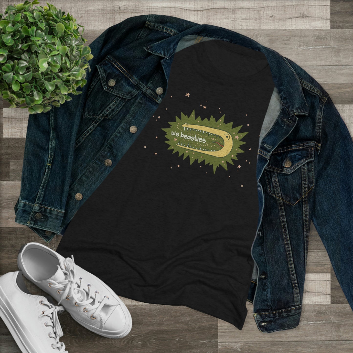 A black t-shirt with the We Beasties logo (a yellow alligator on a green star burst surround by pink stars and the words "We Beasties" in the alligator's mouth.) It's shown with a denim jacket, white sneakers, and a plant against a wood floor.
