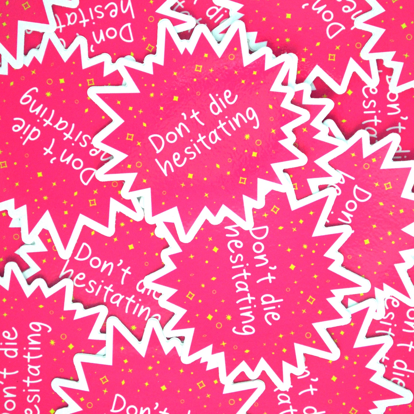 A pile of bright pink star-burst shaped stickers with the words "Don't die hesitating" in white handwriting surrounded by yellow stars and sparkles