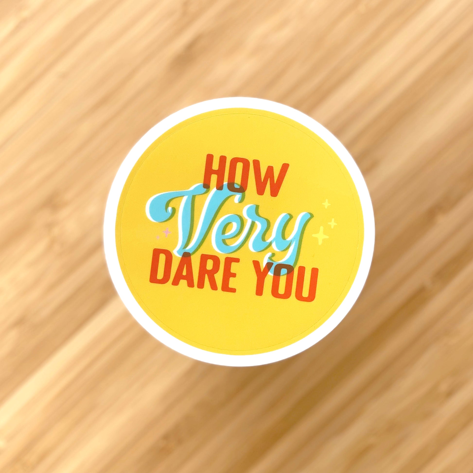 A yellow sticker with orange-red, light blue, and white text that says How Very Dare You. The word "dare" is written in script and the rest of the words are in blocky capital letters. The sticker sits aove a blurred bamboo table top.