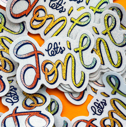A rainbow glitter sticker that says "let's don't" in calligraphy and sits on a ruled background to show it's size.