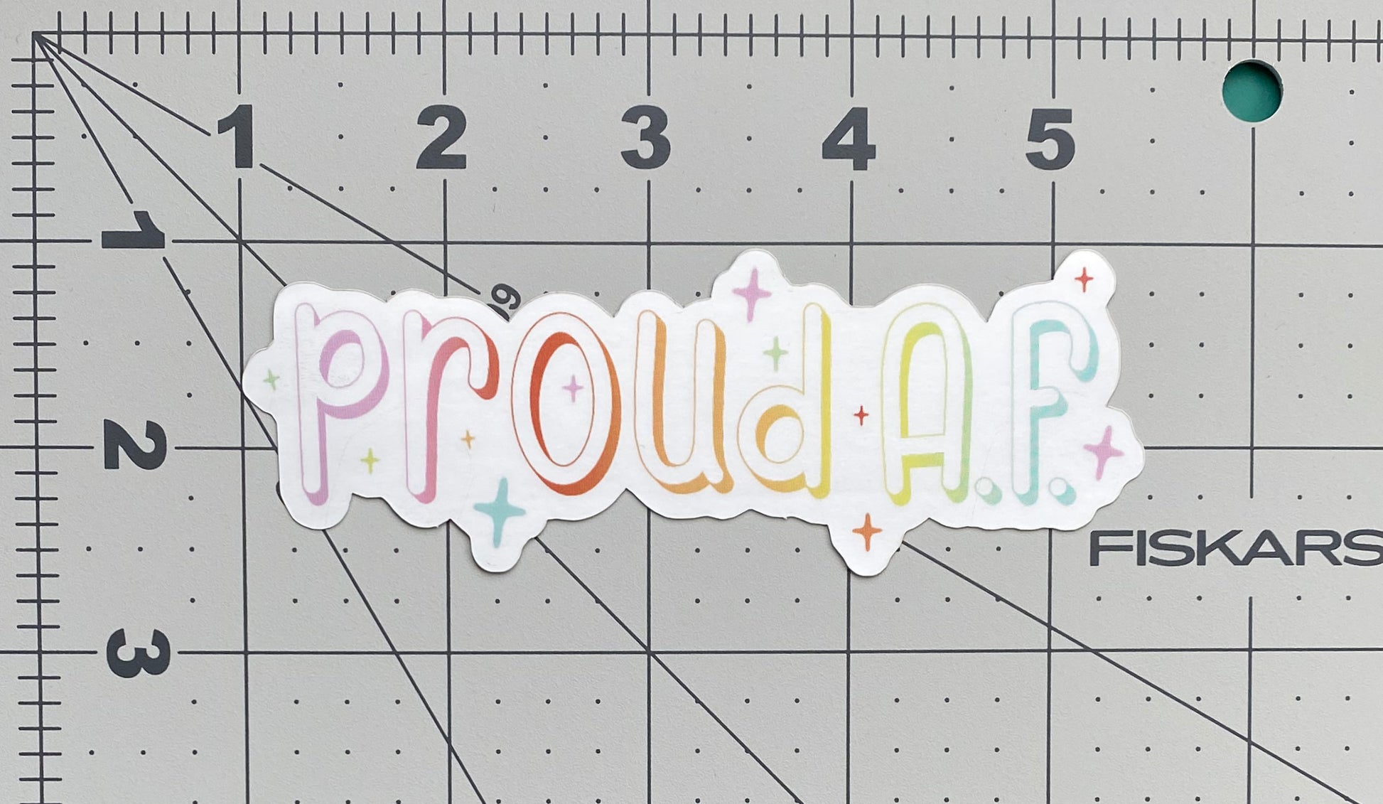 A metallic silver sticker against a ruled background to show its size. The sticker says "proud A.F." in a pastel rainbow gradient and is surrounded by sparkles.