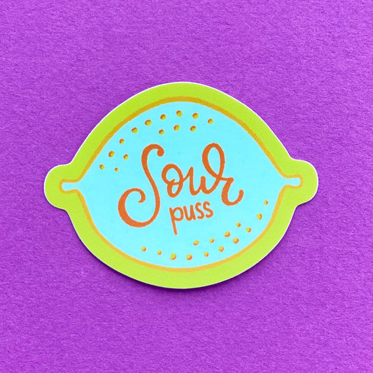 A vinyl sticker with an illustration of a blue lemon and the words "Sour puss" written on it. The sticker has a yellow border and sits on a birght purple paper background.