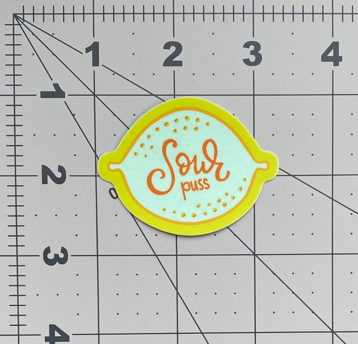 A vinyl sticker with an illustration of a blue lemon and the words "Sour puss" written on it. The sticker has a yellow border and sits on a ruled background to show size.