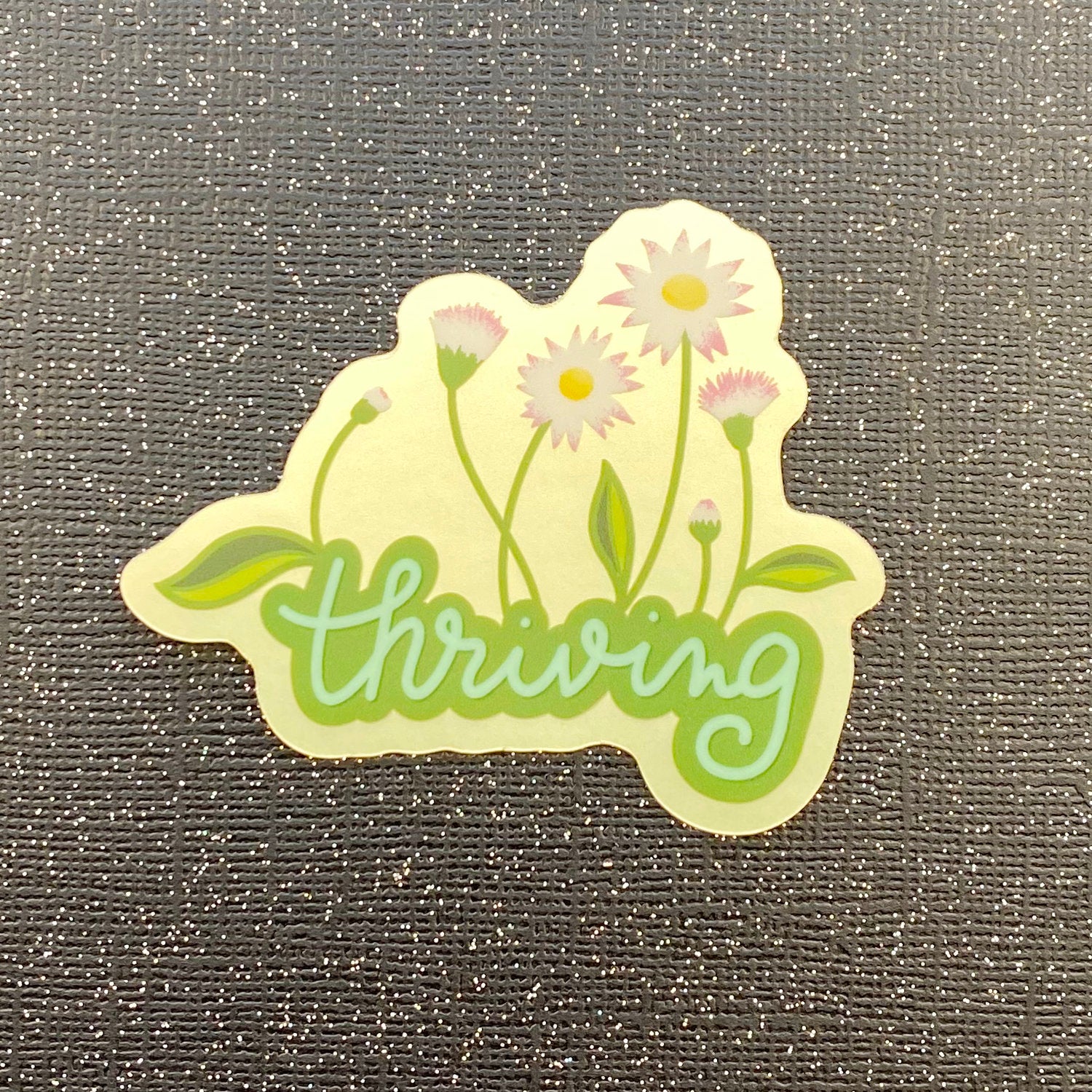 A metallic sticker with the word "thriving" growing white, pink, and yellow flowers on a pale green metallic background. Sticker sticks on black glitter paper.
