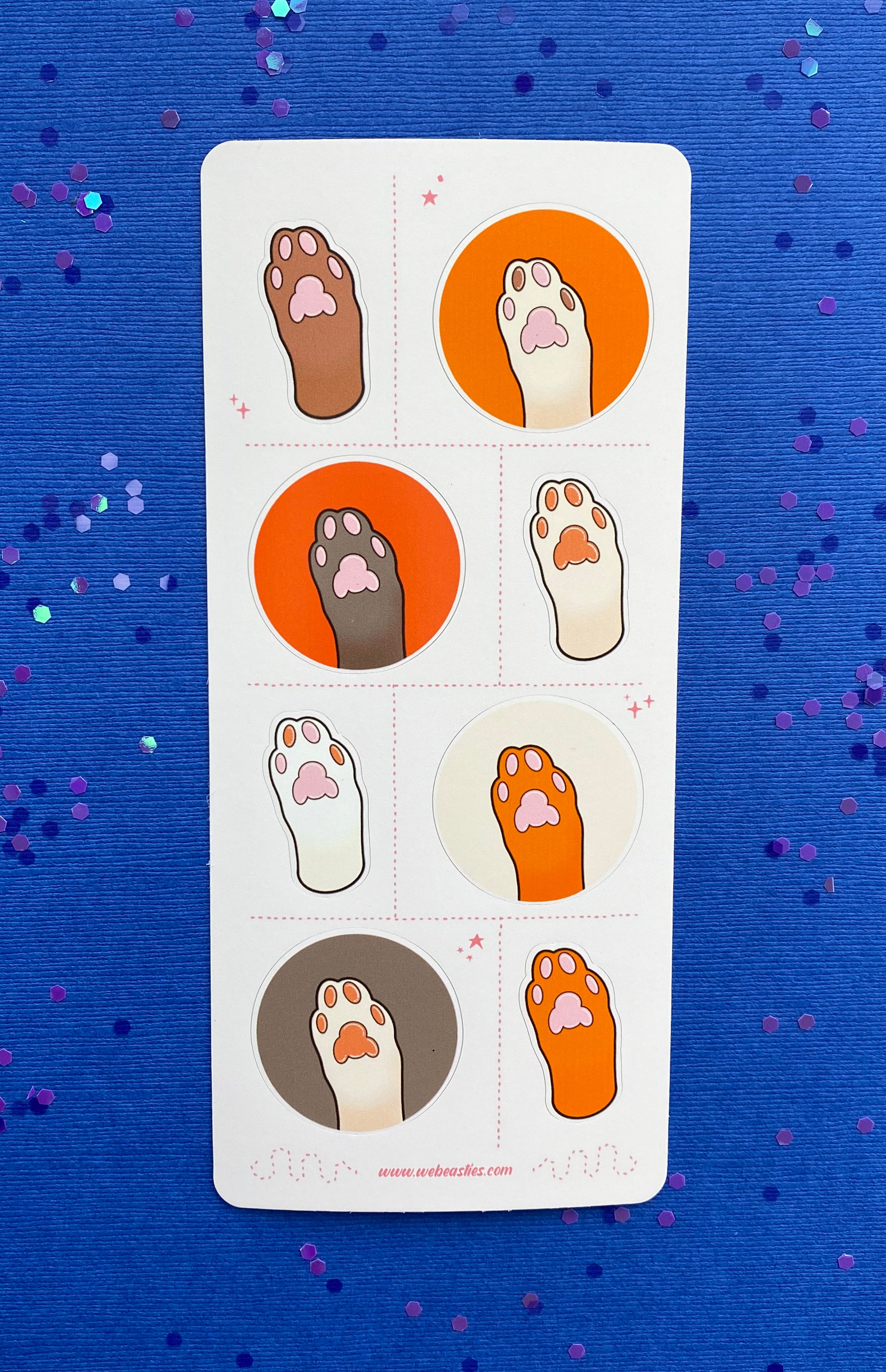 A sticker sheet featuring 8 illustrations of cat's paws and toe pads. The paws are light brown, dark brown, white, and off white. The toe pads are pink, brown, or both. Four paws sit against orange, dark orange, cream or light brown backgrounds and the others are an outline of the paw with no background. The sticker sheet sits against a blue paper background scattered with chunky blue and purple glitter.