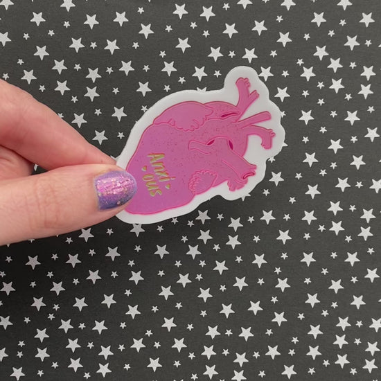 A person with purple nail polish holds a metallic sticker with a realistic human heart illustration and the the word "anxious" flanked by hearts. 