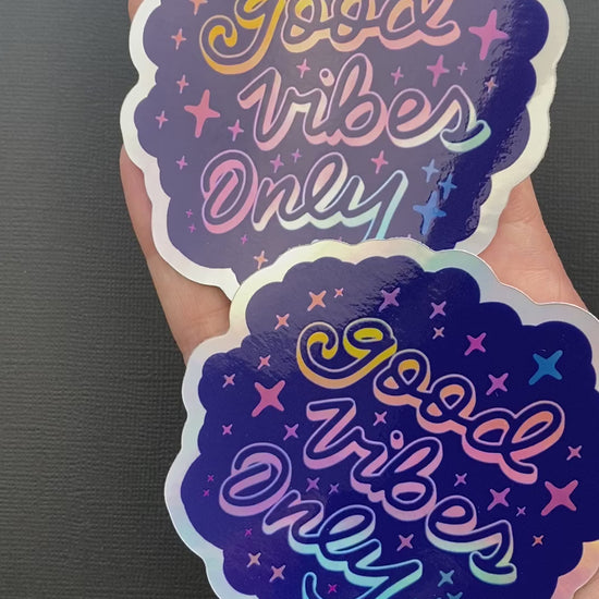 A person's hand holding two holographic "Good Vibes Only" stickers. The hand rotates the stickers to show of their glossy finish and holographic effects.