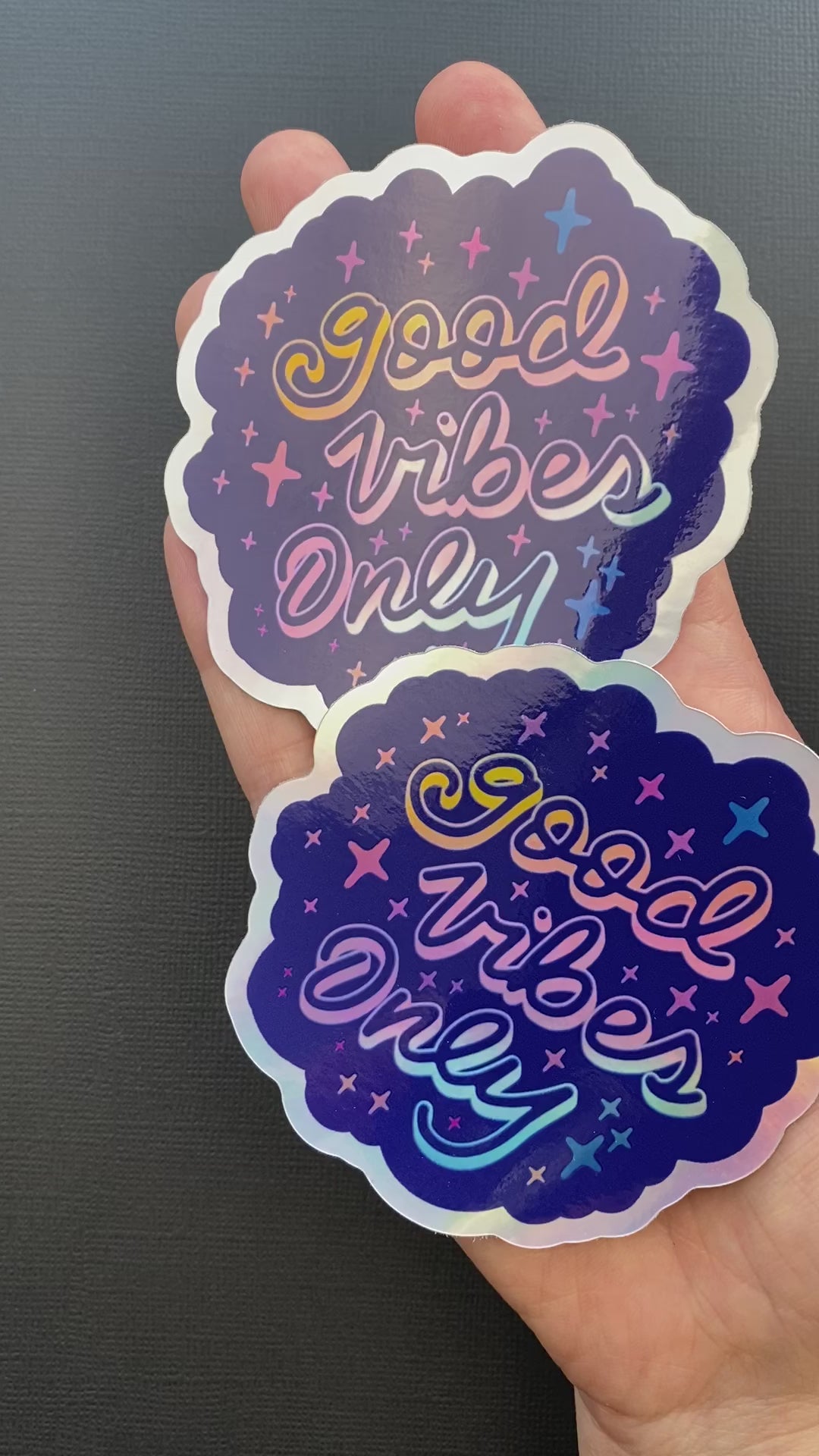 A person's hand holding two holographic "Good Vibes Only" stickers. The hand rotates the stickers to show of their glossy finish and holographic effects.