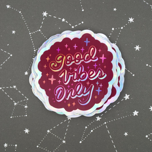 A magenta holographic sticker with the words "good vibes only" written in calligraphy and surrounded by rainbow sparkles. The sticker sits on a black background with constellations on it.