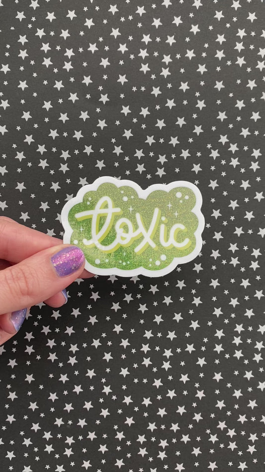 A person with purple nail polish holding a green glitter sticker that says "Toxic" in script. The hand rotates the sticker to show how it catches light. The background is black paper with silver stars.