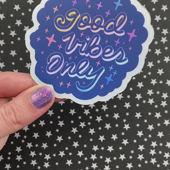 A video of a person holding a blue holographic sticker with the words "good vibes only" written in calligraphy and surrounded by rainbow sparkles. The person rotates the sticker to show its holographic finish.