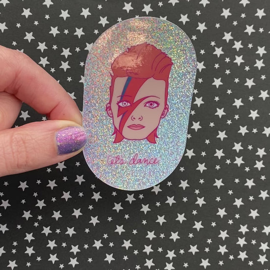 A person with purple nail polish holds a sticker featuring an illustration of David Bowie's head with the phrase "let's dance" on irridescent glitter vinyl. The sticker catches the light as the person turns it.