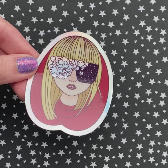 A person with purple nail polish holding an illustrated sticker of Lady Gaga wearing a red hood and black and irridescent glasses against a black background with silver stars