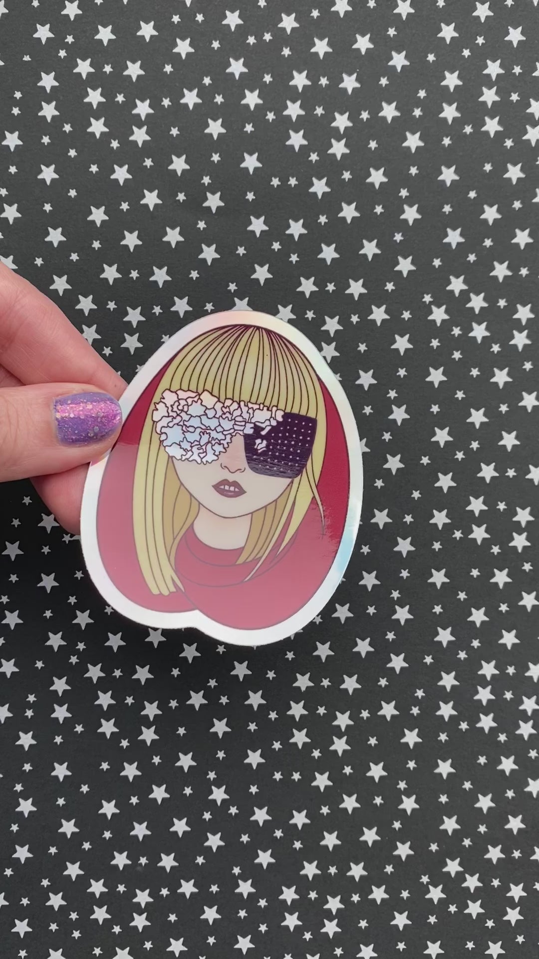A person with purple nail polish holding an illustrated sticker of Lady Gaga wearing a red hood and black and irridescent glasses against a black background with silver stars