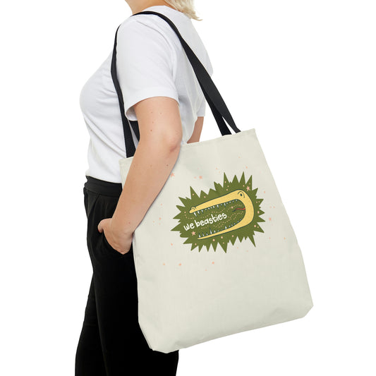 A woman wearing a tote bag slung over her shoulder. The tote has off-white with black straps and is printed with the We Beasties logo (a yellow alligator on a green star burst background surrounded by pink stars and the words "We Beastiest" in its mouth).