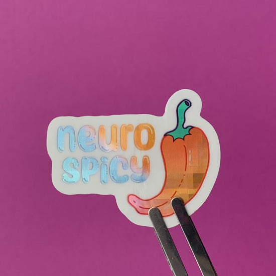 A clip holding a holographic vinyl sticker with an illustration of a pepper and the words "neuro spicy". The holographic material is a blue to purple to orange gradient pattern with a plaid effect. The sticker rotates to show the holographic effect in different light.