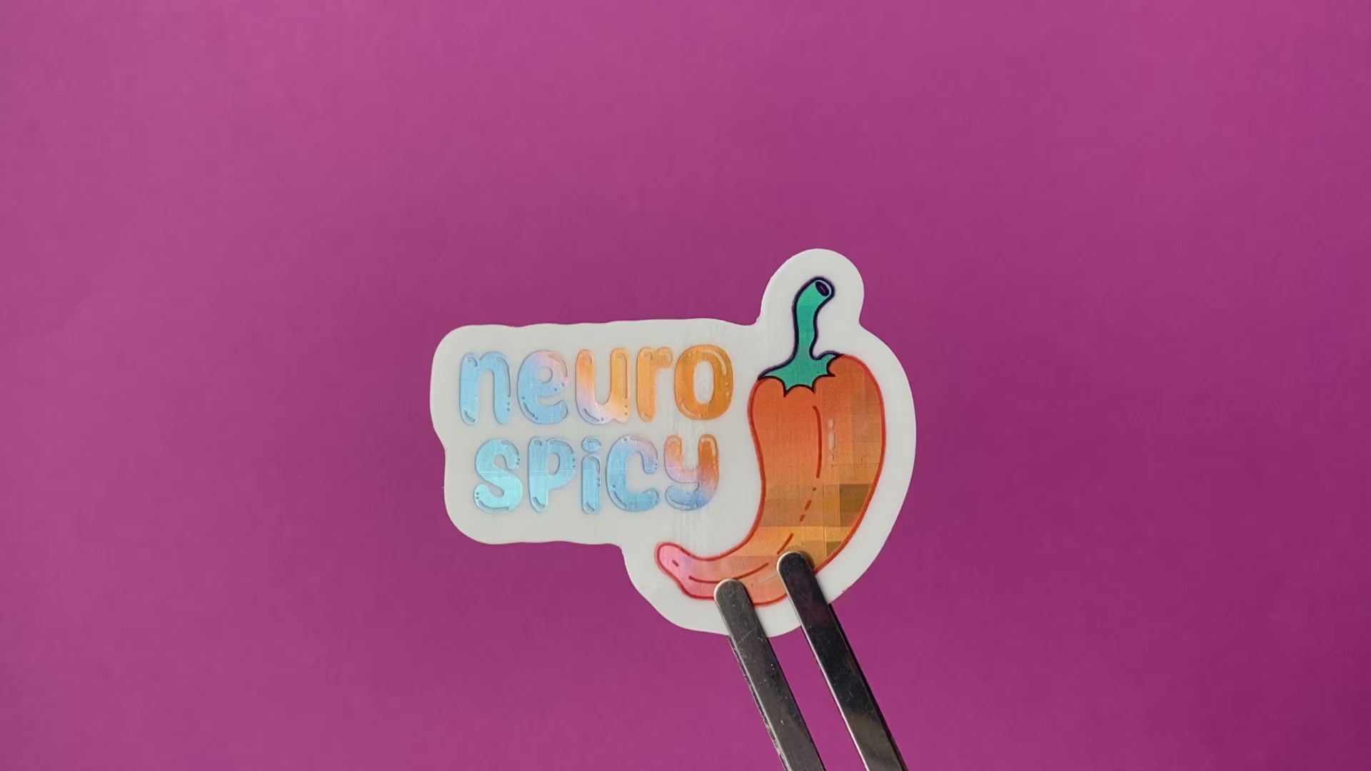 A clip holding a holographic vinyl sticker with an illustration of a pepper and the words "neuro spicy". The holographic material is a blue to purple to orange gradient pattern with a plaid effect. The sticker rotates to show the holographic effect in different light.