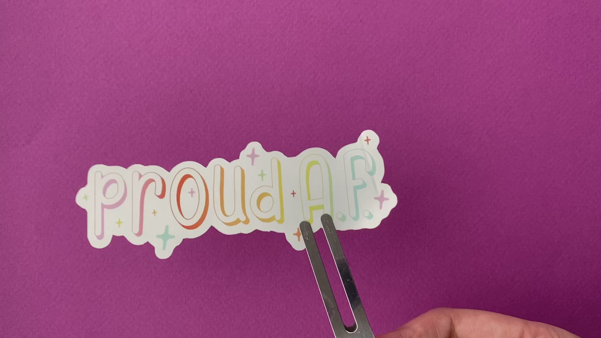 A clip holds a metallic silver sticker against a bright purple paper background. The sticker says "proud A.F." in a pastel rainbow gradient and is surrounded by sparkles. As the sticker rotates, the metallic surface reflects lightly at different angles.