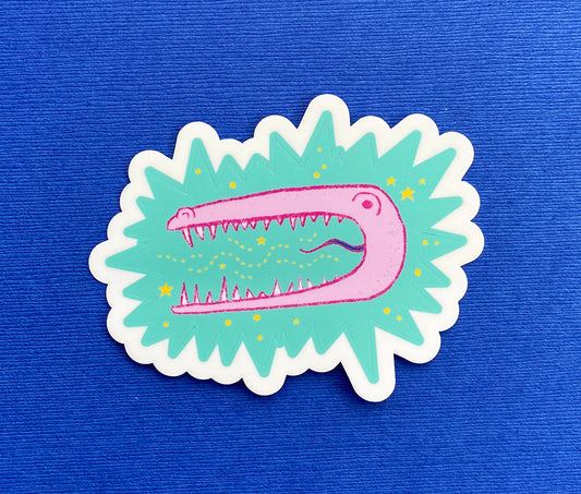 A child-like illustration of a pink alligator with sharp teeth. It's surrounded by yellow stars and sits on a turquoise star burst. The sticker is kiss cut.