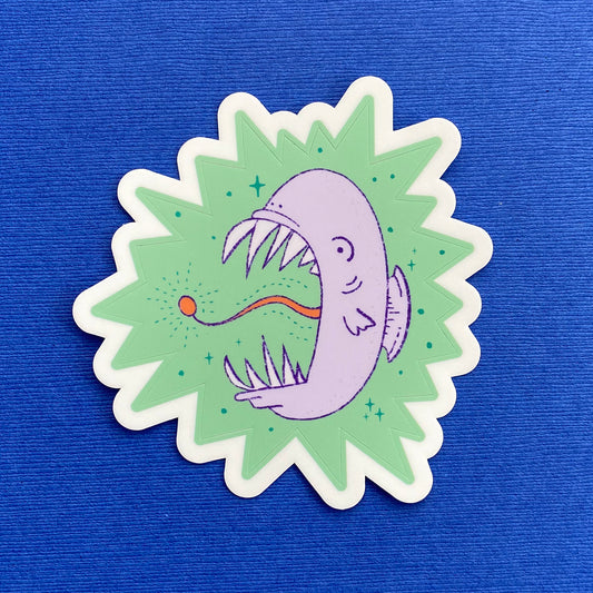 A child-like illustration of a purple angler fish with sharp teeth. It's surrounded by blue-green stars and sits on a mint green star burst. The sticker is kiss cut.