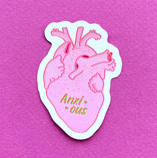 A metallic sticker with an illustration of a realistic human heart and the the word "anxious" flanked by hearts. The sticker sits on a ruler to show its size.