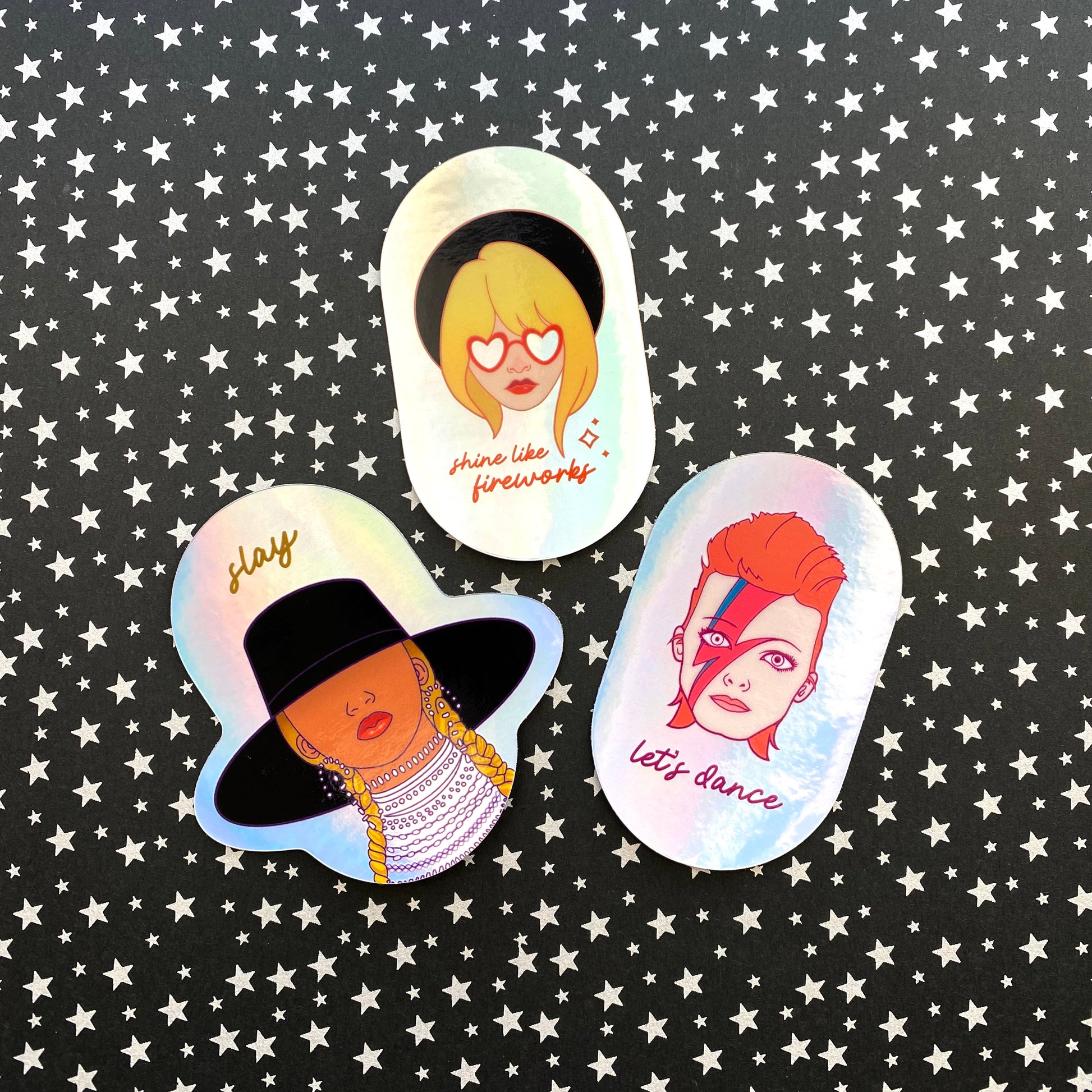 A photo of three holographic stickers illustrated with portraits of Beyoncé, Taylor Swift and David Bowie. The sticker sits on a black and silver star background.