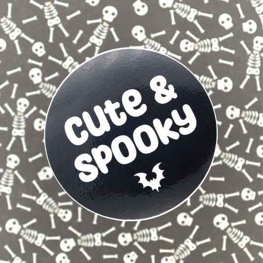 A black sticker with a small bat and the words "cute and spooky" in white ink. The sticker sits against a black and white background featuring cute skelletons