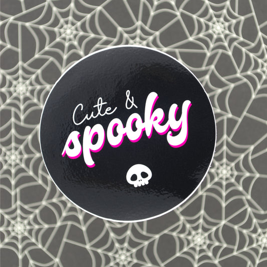 A glossy black vinyl sticker with a skull and the words "Cute & Spooky" and spooky has a pink drop shadow. The sticker sits on a black and white spiderweb background