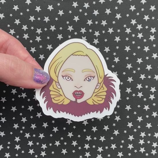 A person with purple nail polish holding an illustrated sticker of Lady Gaga from American Horror Story to show it's holographic material. The background is black paper with silver stars.