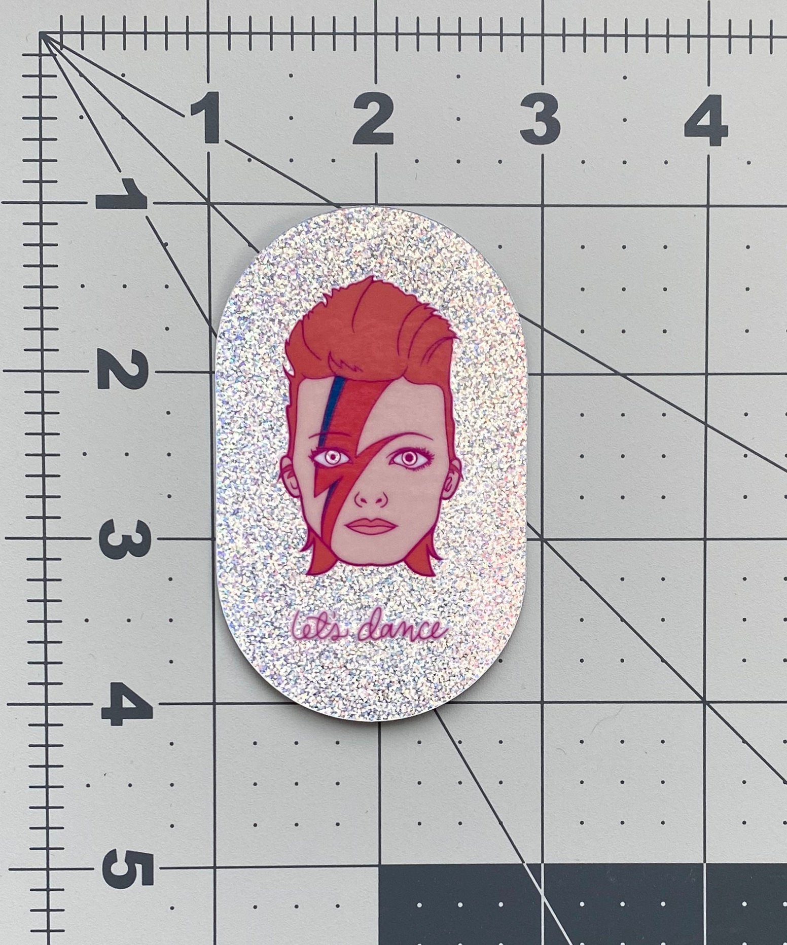 An illustration of David Bowie's head with the phrase "let's dance" on irridescent glitter vinyl. The sticker sits on a ruler mat to show the sticker's size.