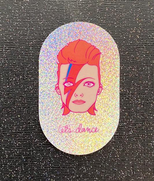 An illustration of David Bowie's head with the phrase "let's dance" on irridescent glitter vinyl. The sticker sits on black paper with silver glitter.