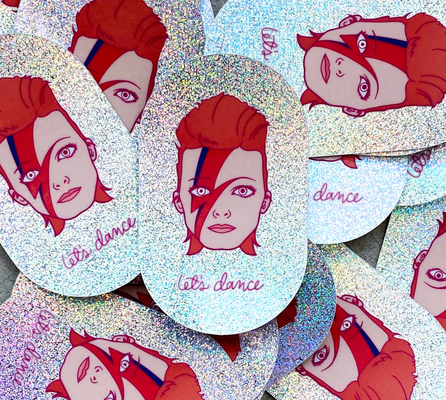 A pile of stickers featuring an illustration of David Bowie's head with the phrase "let's dance" on irridescent glitter vinyl. 