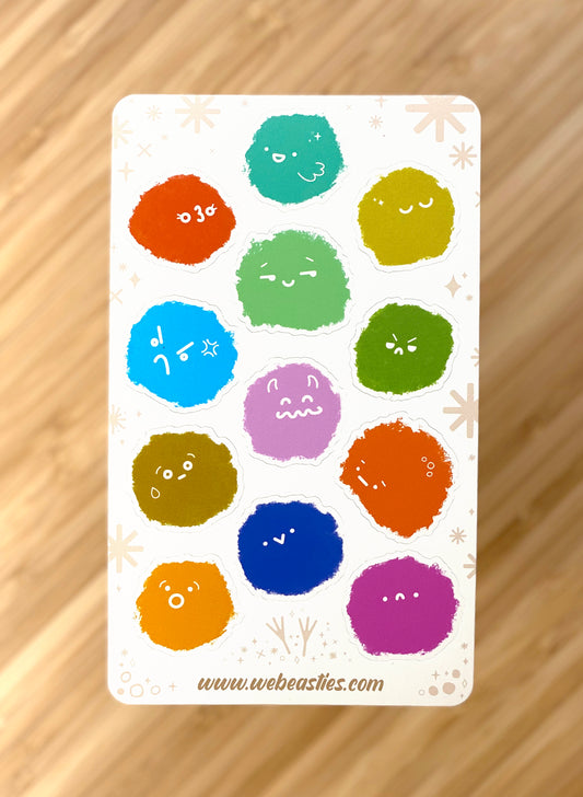 A photo of a sticker sheet featuring 12 fuzzie textured blobs with a variety of expressions. The sheet sit above a blurred bamboo background.