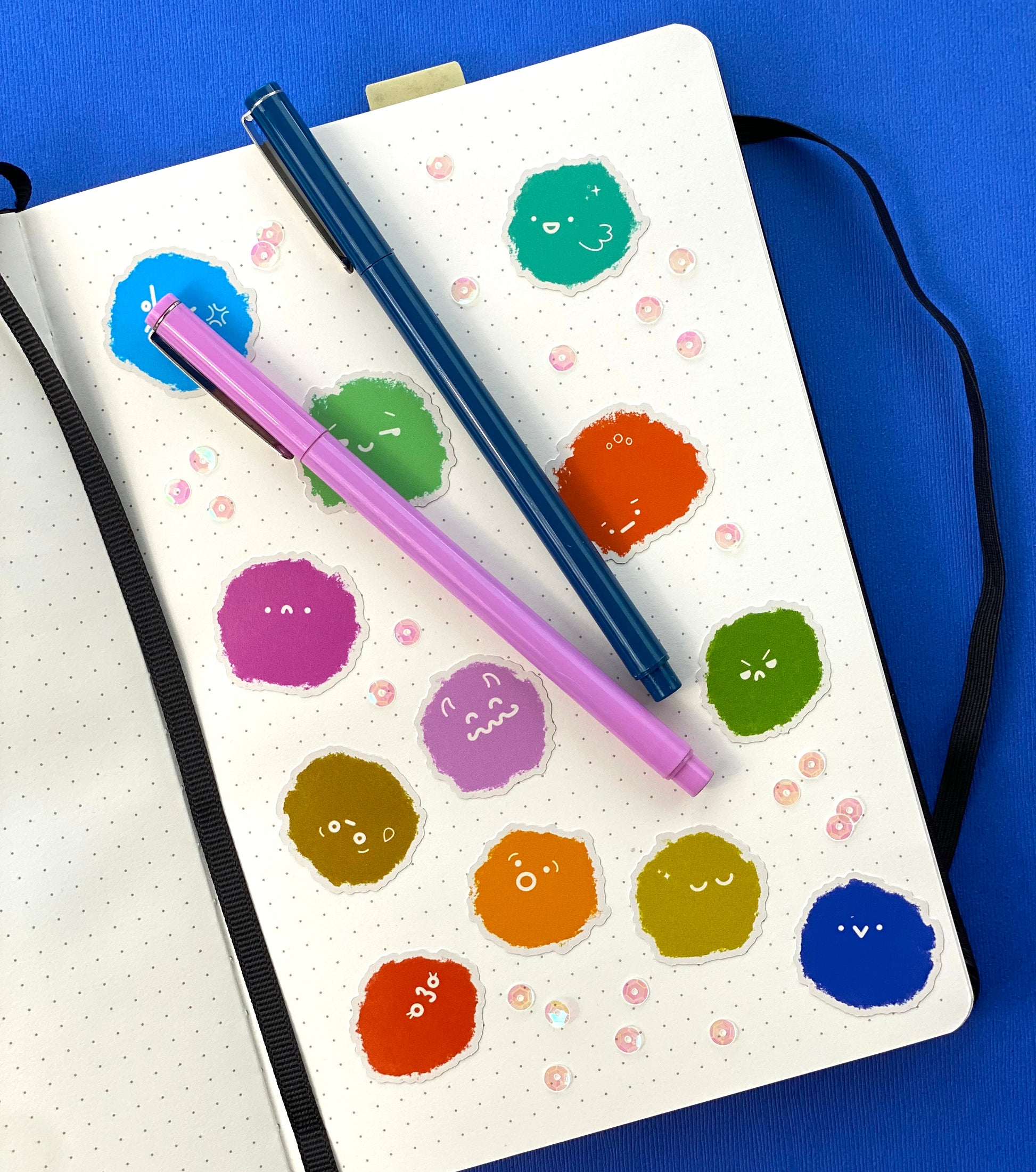 A photo of a journal page covered in sequins and stickers of 12 fuzzie textured blobs with a variety of expressions. Two pens, one blue and one pink, sit on the journal page. The journal sits on a blue background.