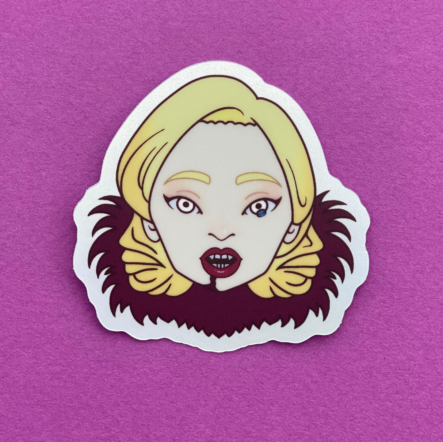 A sticker featuring an illustration of Lady Gaga's head as she appears in American Horror Story. She has a tear in one eye, vampire fangs, red lipstick, and a red fur collar. Her hair is coiffed in a retro 1940's inspired style.