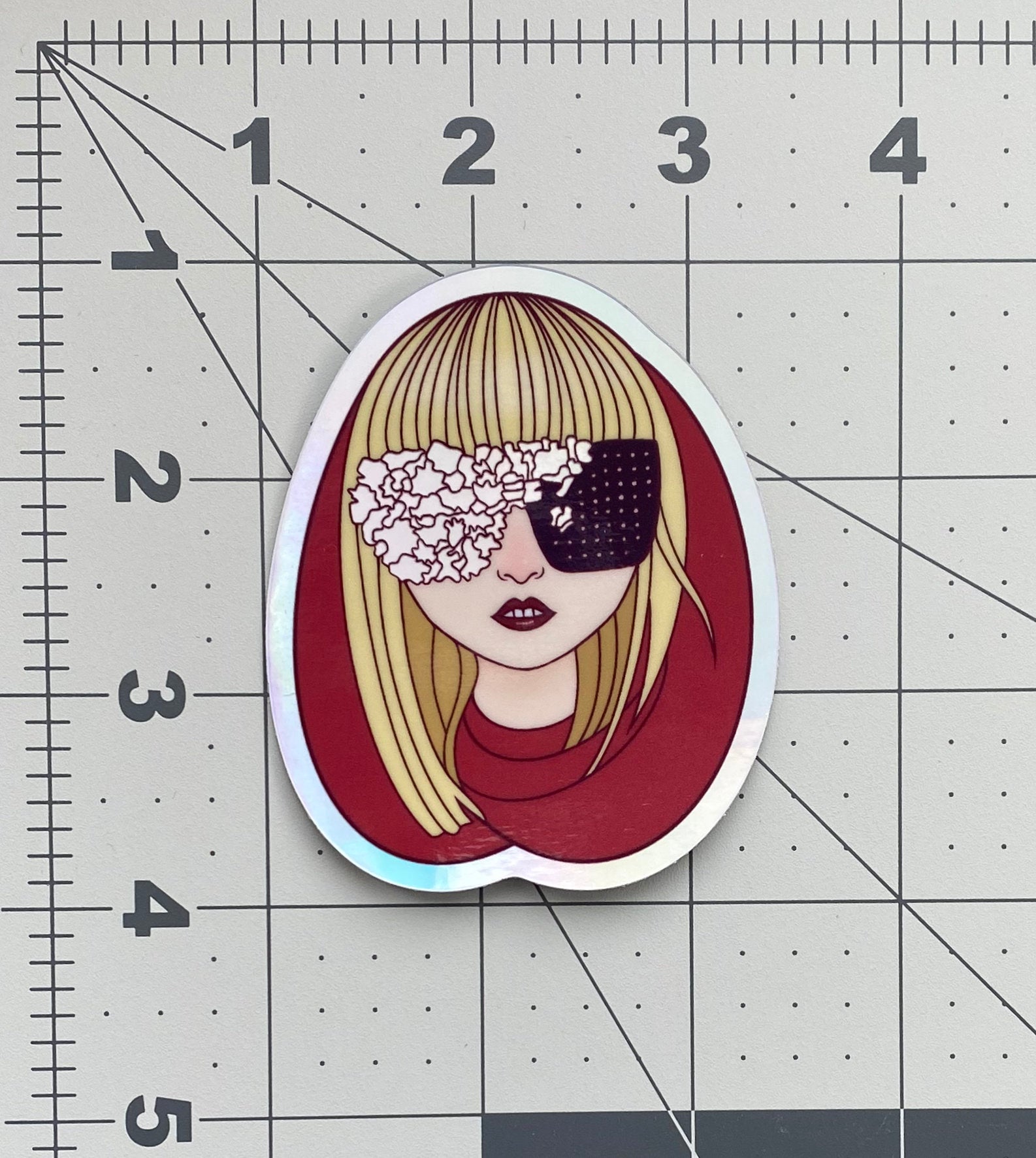 Illustrated sticker of Lady Gaga wearing a red hood and black and irridescent glasses against a ruled background to show sticker size