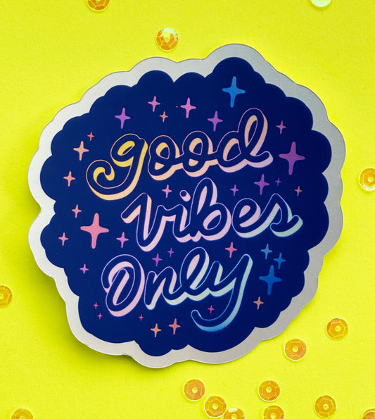 A blue holographic sticker with the words "good vibes only" written in calligraphy and surrounded by rainbow sparkles. The sticker sits on a neon yellow background scattered with irridescent sequins.