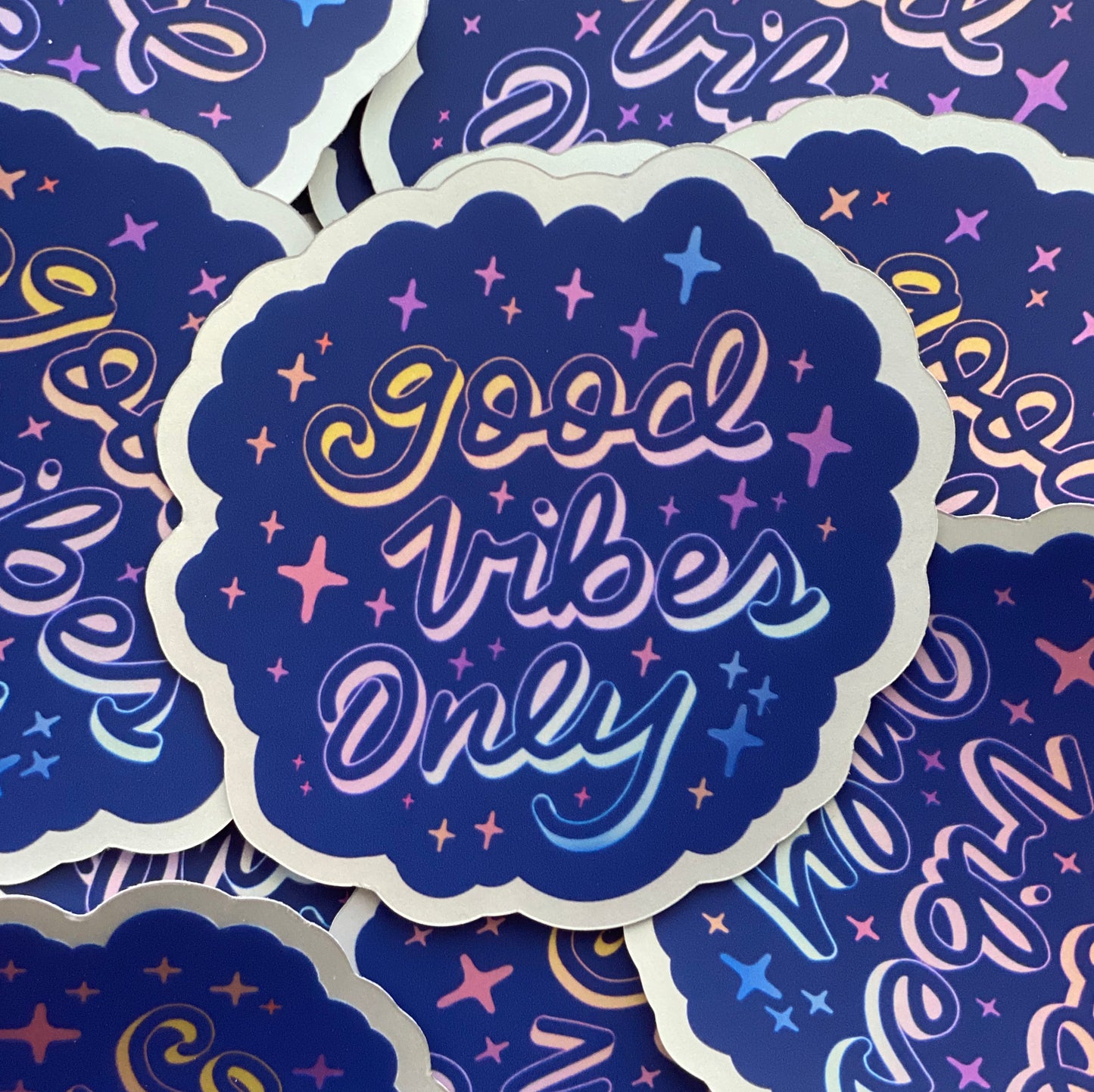 A pile of blue holographic stickers with the words "good vibes only" written in calligraphy and surrounded by rainbow sparkles. 