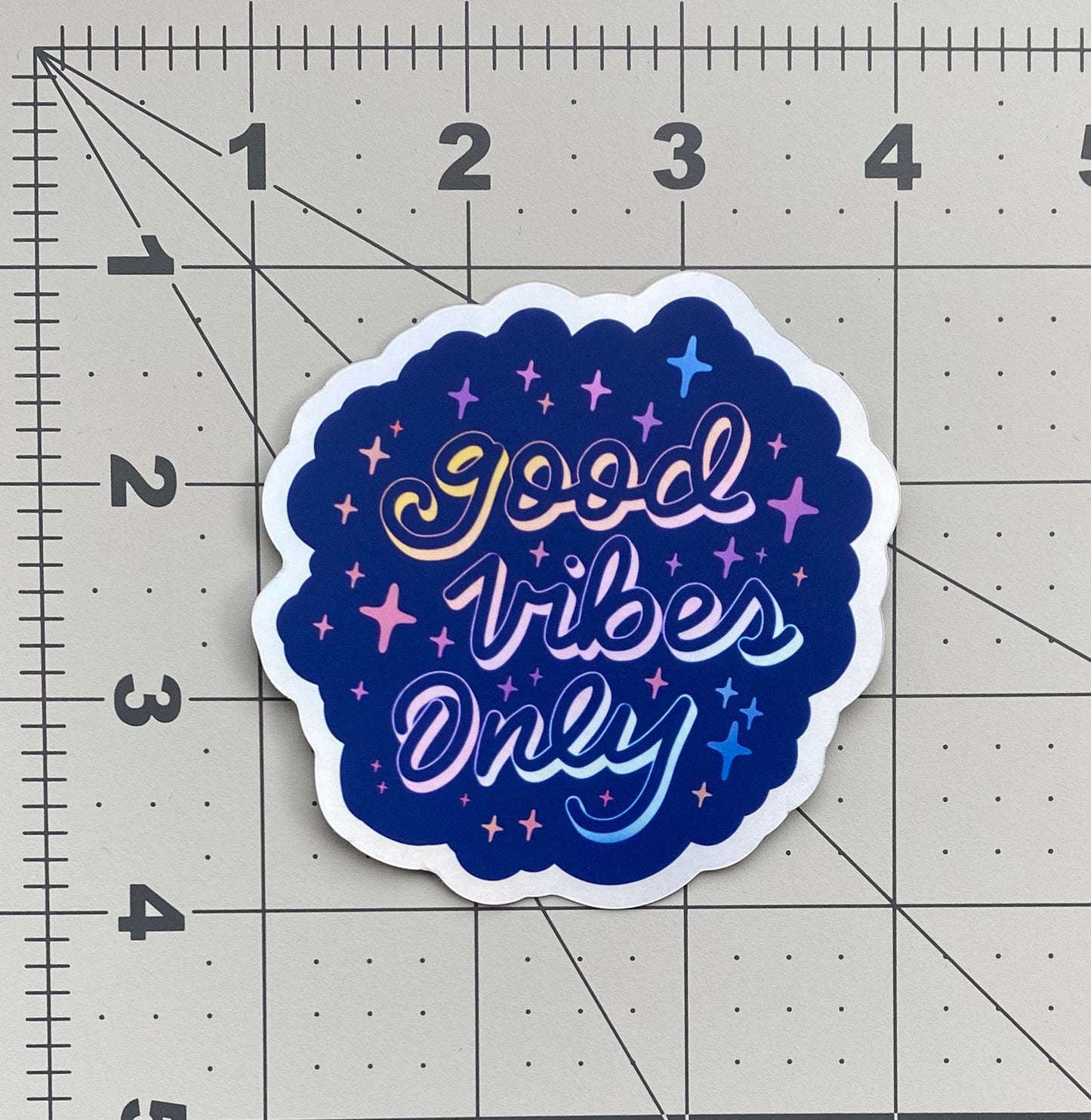 A blue holographic sticker with the words "good vibes only" written in calligraphy and surrounded by rainbow sparkles. The stickers sits on a ruled background to show size.