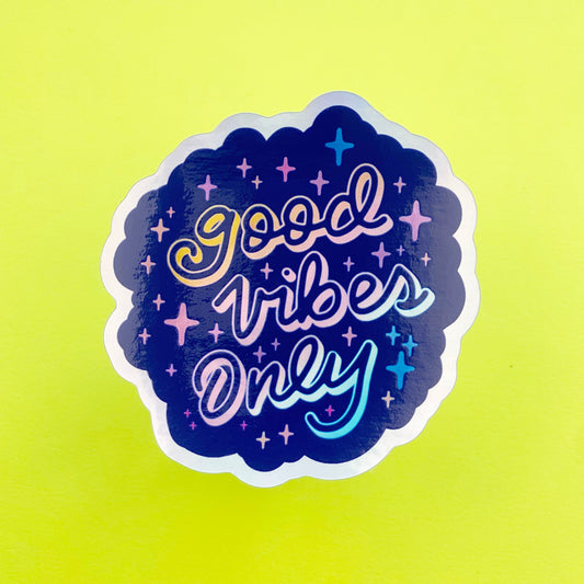 A blue holographic sticker with the words "good vibes only" written in calligraphy and surrounded by rainbow sparkles. The sticker sits on a neon yellow background.